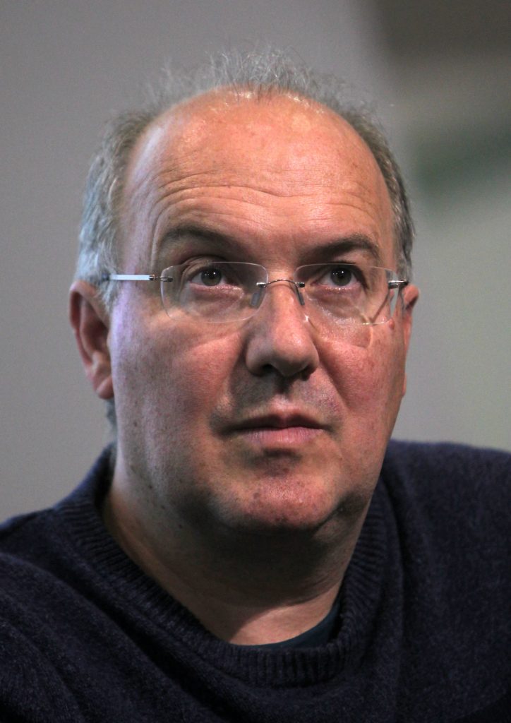 Alain Damasio en gros plan, lunettes fines et pull sombre, sur un fond gris