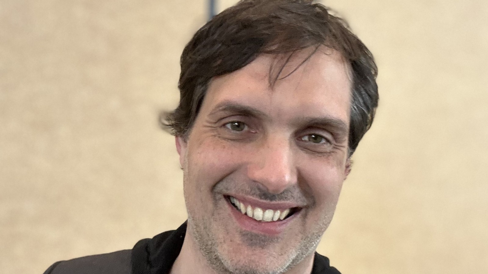 Olivier Derivière, cheveux bruns en bataille et barbe de quelques jours, sourit devant un mur beige uni, en veste grise sur un sweat noir à capuche