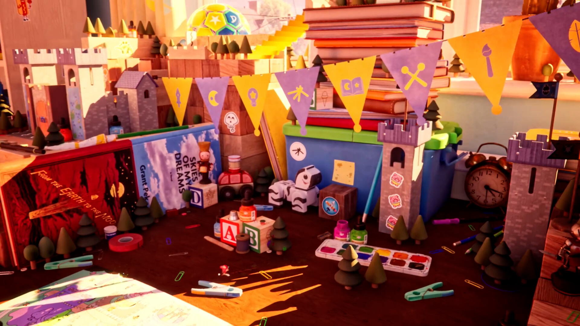 Un bureau d'enfant vu au ras du plateau, avec un château de carton, des fanions jaunes et violets, des piles de livres, des cubes en bois, une palette d'aquarelle et un réveil