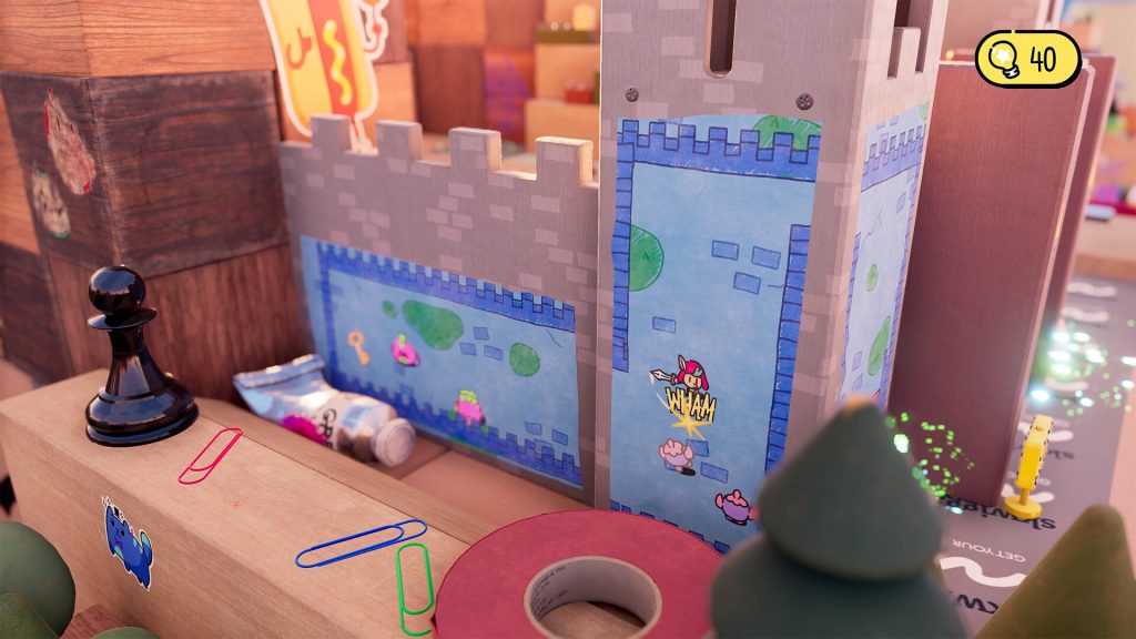 Un dessin plat collé sur la paroi d'un château de carton, où un petit personnage frappe un ennemi, entre une pièce d'échecs noire et des trombones colorés posés sur le bureau