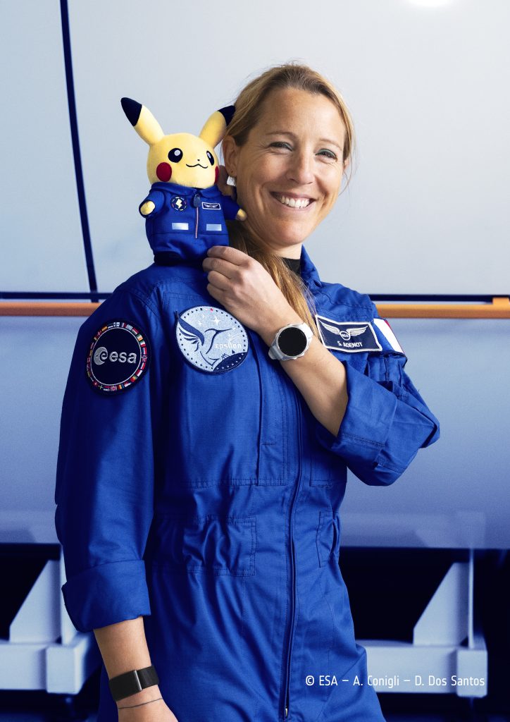 Sophie Adenot, en combinaison de vol bleue de l'ESA, sourit en tenant contre son épaule une peluche Pikachu vêtue d'une combinaison bleue.