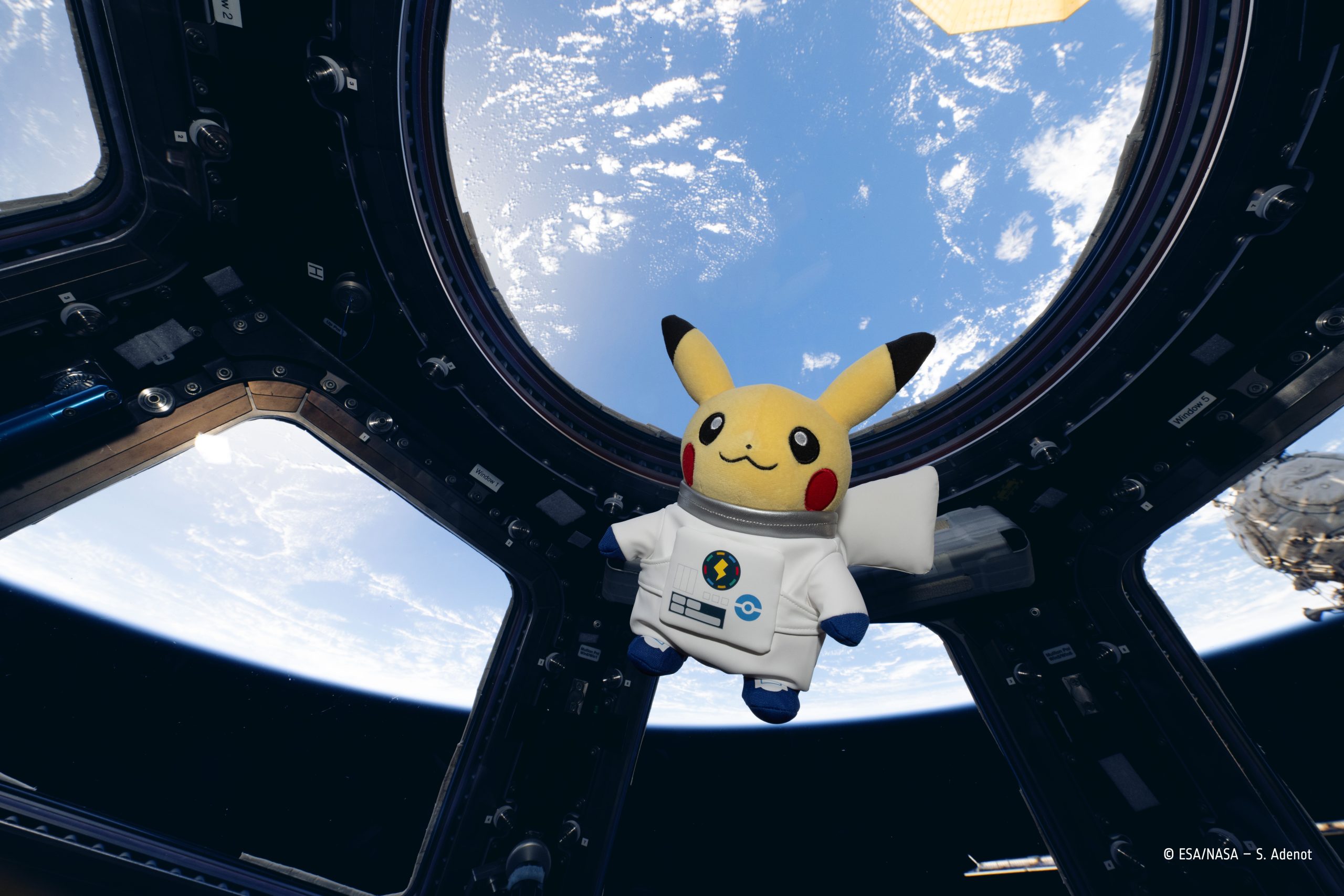 Une peluche Pikachu en combinaison spatiale blanche flotte devant les hublots de la Cupola, la Terre bleue et nuageuse derrière elle.