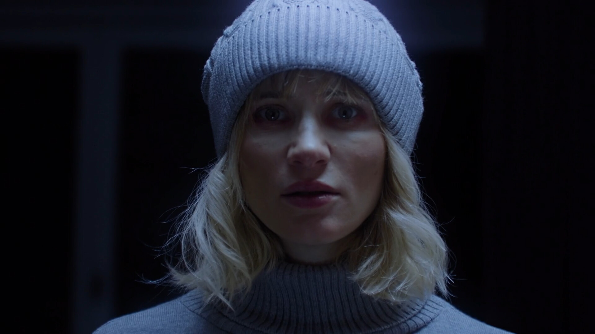 Le visage filmé d'une jeune femme en bonnet de laine claire, éclairé de face dans une pièce sombre