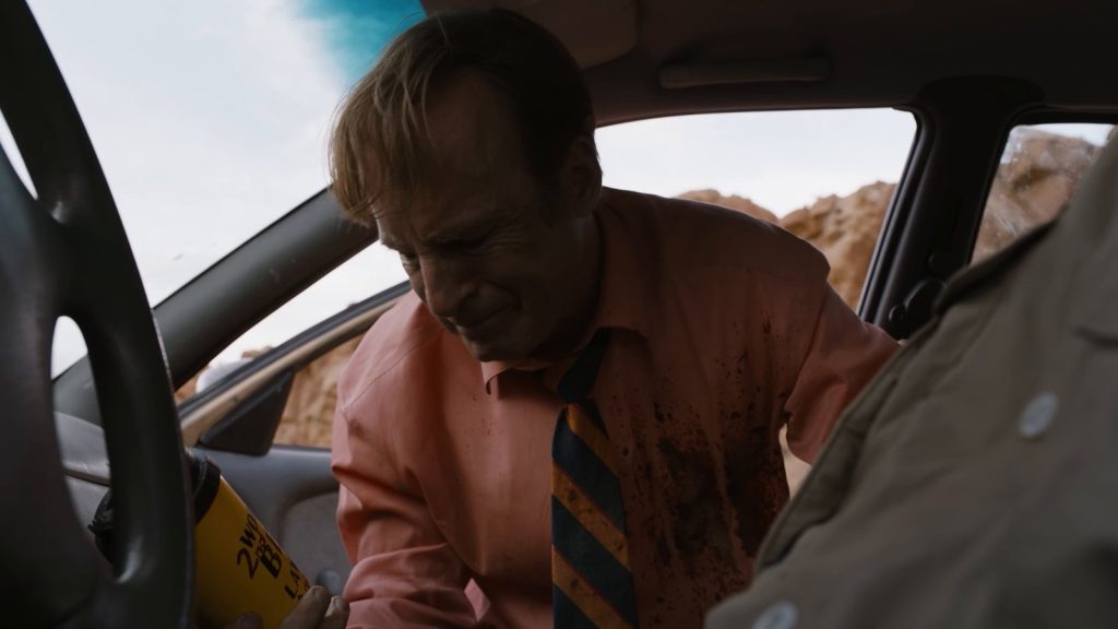 Jimmy, en chemise rose tachée de sang et cravate rayée, tient un gobelet jaune en sortant d’une voiture, devant des rochers ocre.
