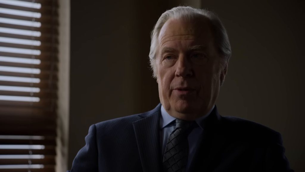 Chuck McGill (Michael McKean), en costume bleu marine, témoigne à la barre, éclairé par une fenêtre à stores.