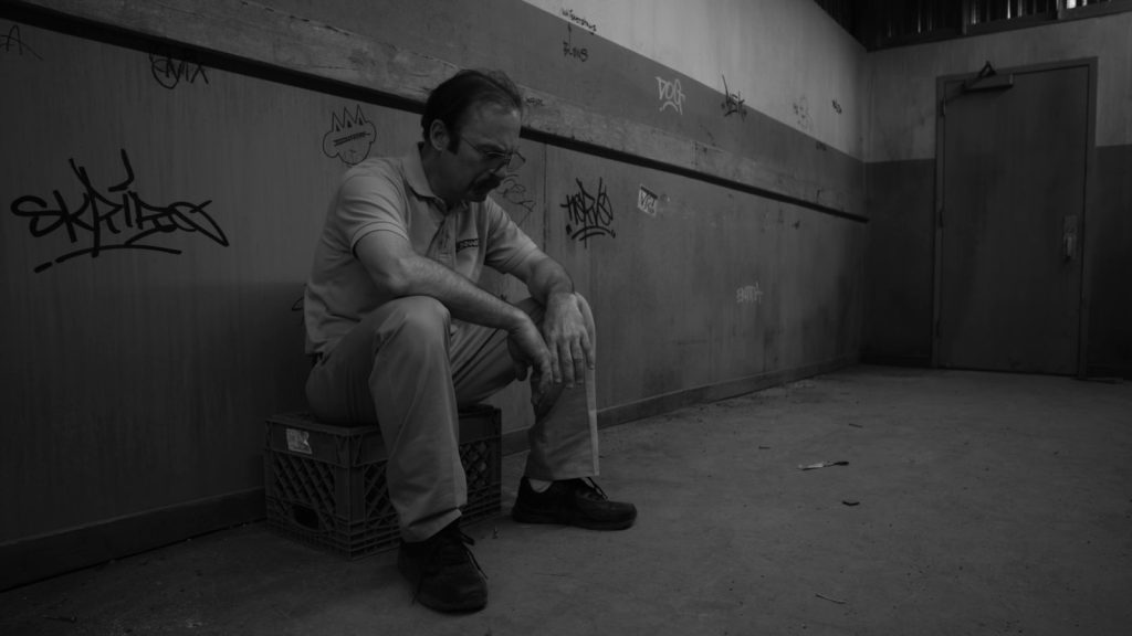 En noir et blanc, Gene Takavic (Bob Odenkirk), en polo, assis tête baissée sur une caisse en plastique dans un couloir de service couvert de tags.
