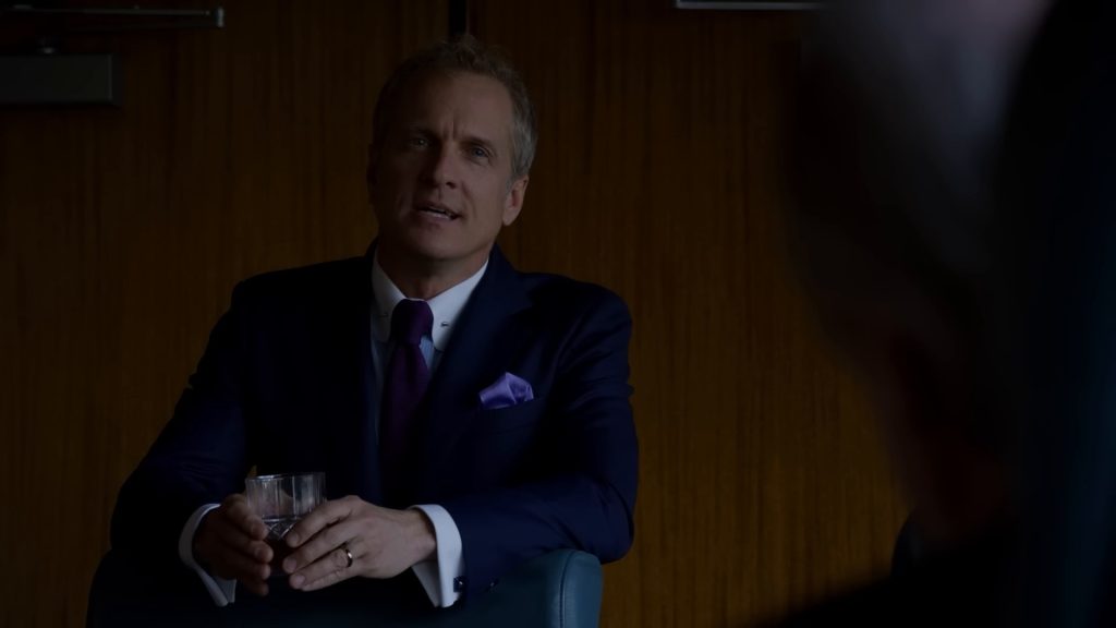 Howard Hamlin (Patrick Fabian), en costume bleu marine, cravate et pochette violettes, un verre à la main dans la salle de réunion de HHM.