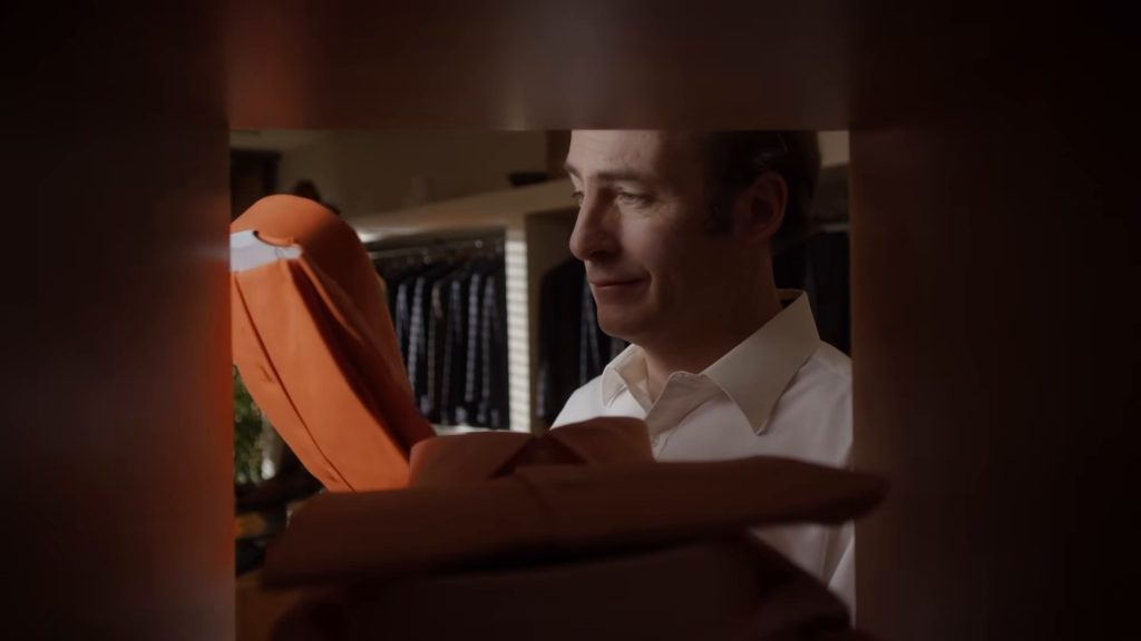 Jimmy McGill (Bob Odenkirk), en chemise blanche, tient une chemise orange vif entre deux rayonnages chez un tailleur.