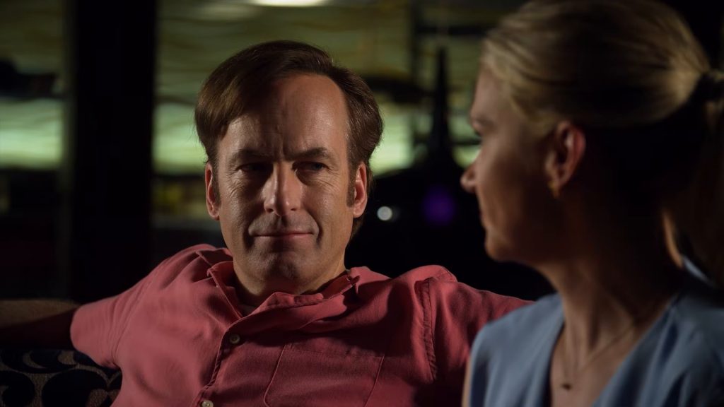 Jimmy McGill, en polo rose saumon, sourit à côté de Kim Wexler, en haut bleu clair, dans un bar de nuit.
