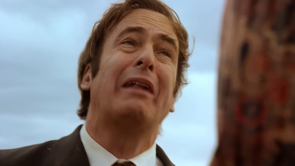 Gros plan sur Jimmy McGill (Bob Odenkirk) en veste brune, le visage crispé, sous un ciel bleu ; à droite, flou, le bord d’une chemise à motifs rouges.
