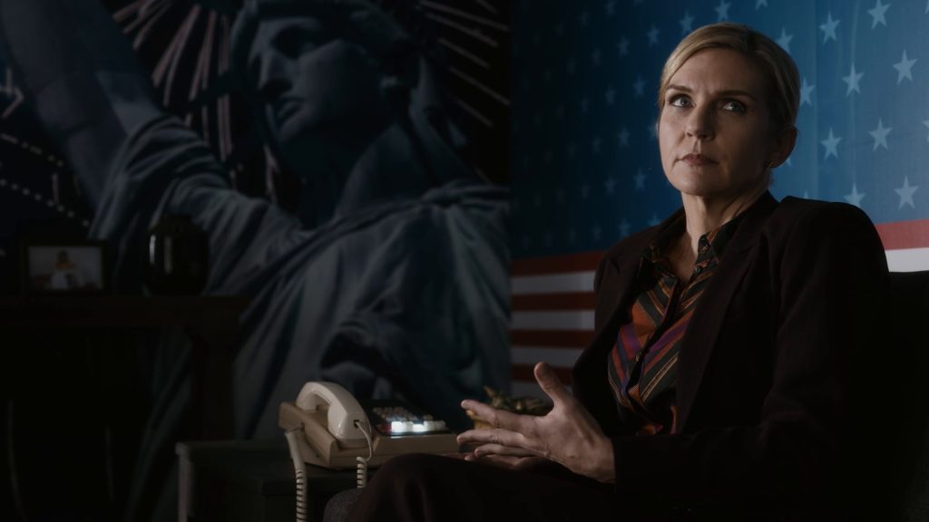 Kim Wexler, en veste sombre et chemisier à rayures chaudes, assise devant un mur peint de la statue de la Liberté et du drapeau américain.