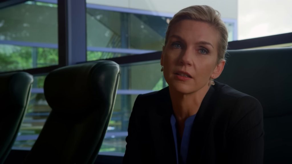 Kim Wexler (Rhea Seehorn), en tailleur noir et chemisier bleu, dans une salle de réunion aux fauteuils de cuir vert.
