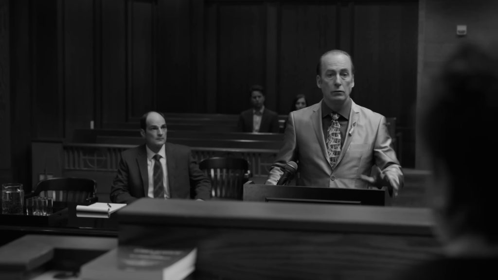 En noir et blanc, Jimmy McGill, en costume clair et cravate à motifs, debout au pupitre d'un tribunal fédéral, un homme assis derrière lui.