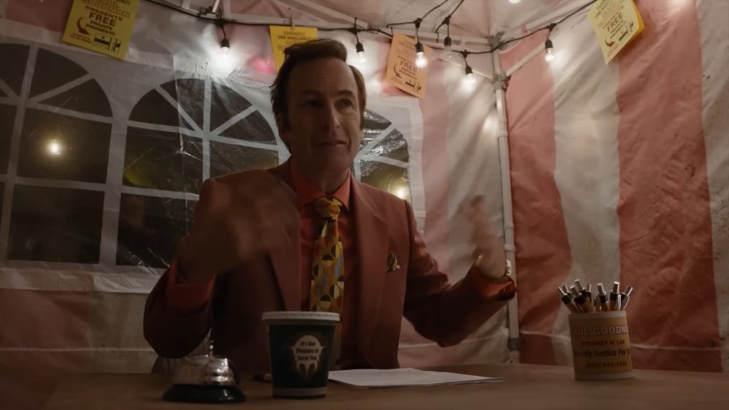 Saul Goodman, en veste rouge corail, chemise orange et cravate jaune à motifs, assis derrière une table sous une tente rayée de rose et de blanc, décorée de guirlandes lumineuses.