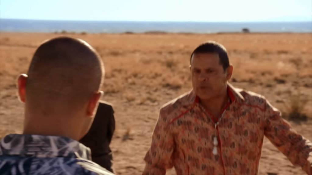 Tuco Salamanca (Raymond Cruz), en chemise à motifs rouge saumon, s’avance dans un désert d’herbes sèches ; au premier plan, de dos, un homme au crâne rasé en chemise à motifs bleus et blancs.