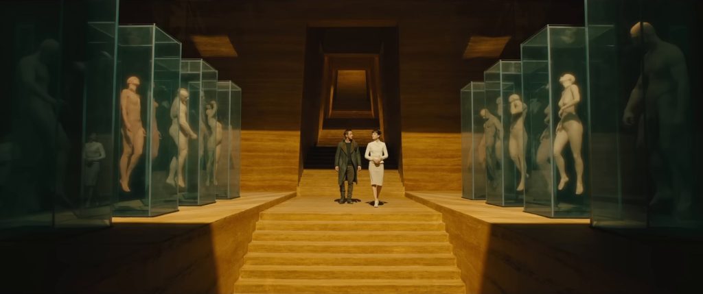 Un homme et une femme descendent un escalier dans un hall doré, bordé de vitrines où se dressent des statues de corps nus, dans Blade Runner 2049.