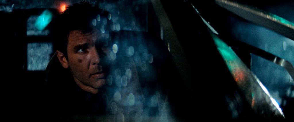 Rick Deckard, joué par Harrison Ford, regarde par la vitre de sa voiture volante, couverte de gouttes de pluie, dans la nuit.