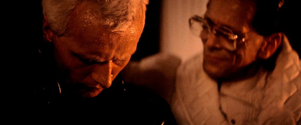 Roy Batty, cheveux blond platine, penché vers Eldon Tyrell, un homme aux grosses lunettes en robe de chambre blanche qui lui sourit, dans une lumière chaude.