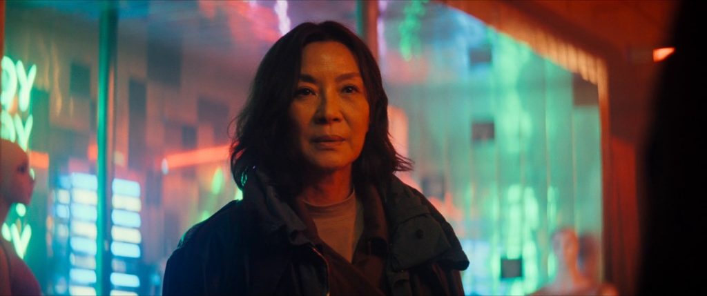 Michelle Yeoh, cheveux mi-longs et veste sombre, devant une vitrine éclairée de néons verts et rouges, dans Blade Runner 2099.