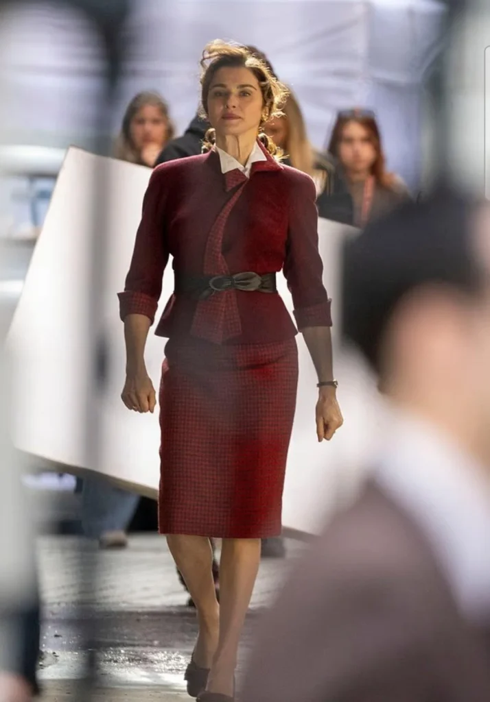 Rachel Weisz en tailleur bordeaux passe devant des panneaux blancs de tournage, photographiée de loin.