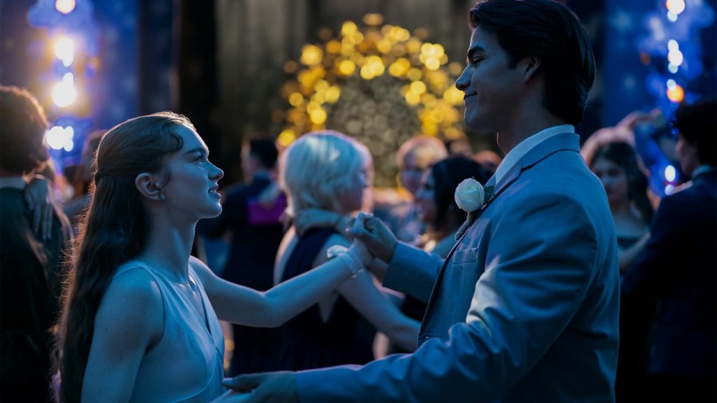 Une adolescente en robe bleu pâle danse face à un garçon en costume clair, main dans la main, sous des guirlandes lumineuses et au milieu des autres couples.
