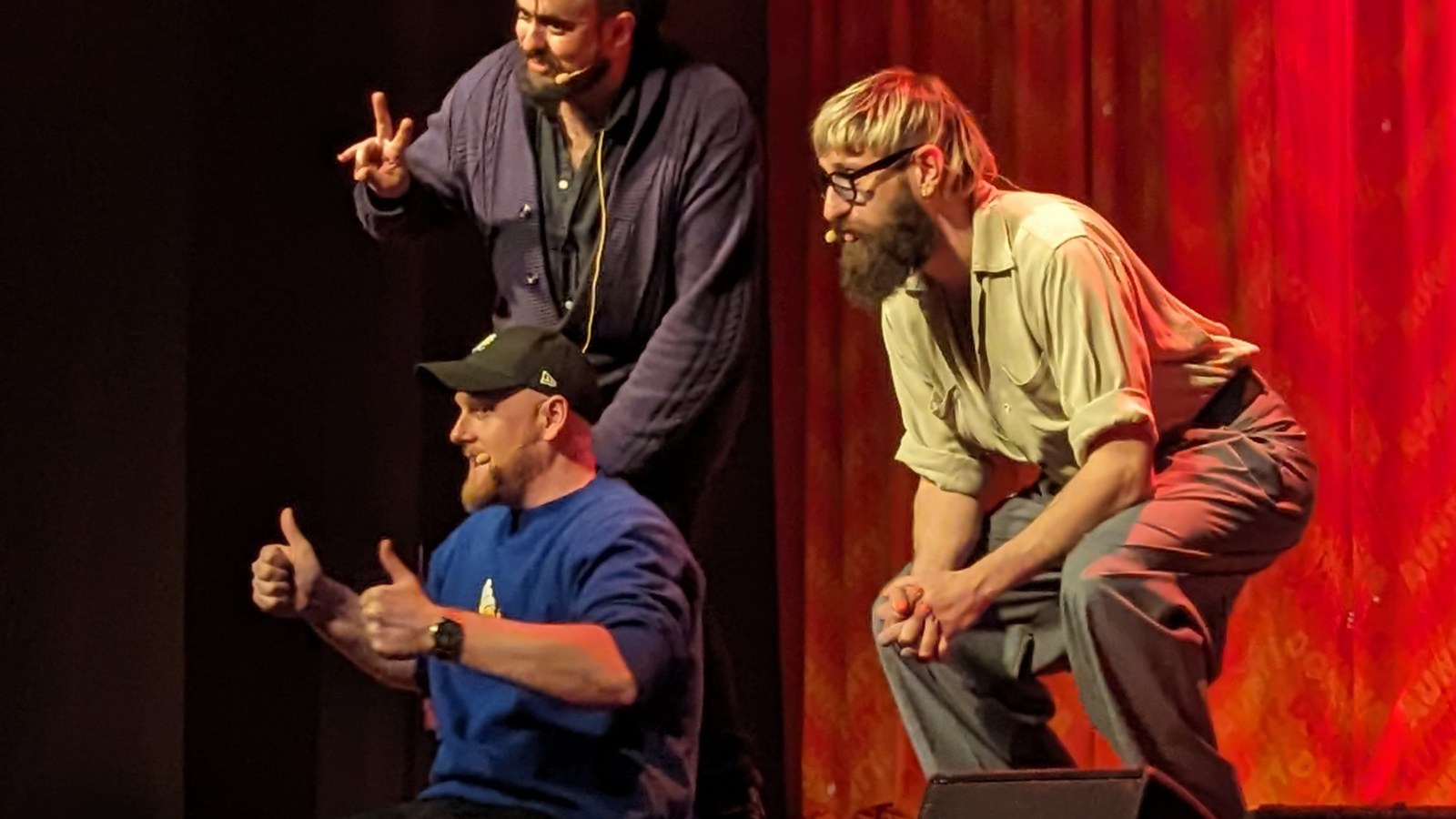 Les trois membres d'Aunty Donna sur une scène de théâtre devant un rideau rouge : l'un pointe du doigt, un autre lève les pouces, le troisième se penche en avant.