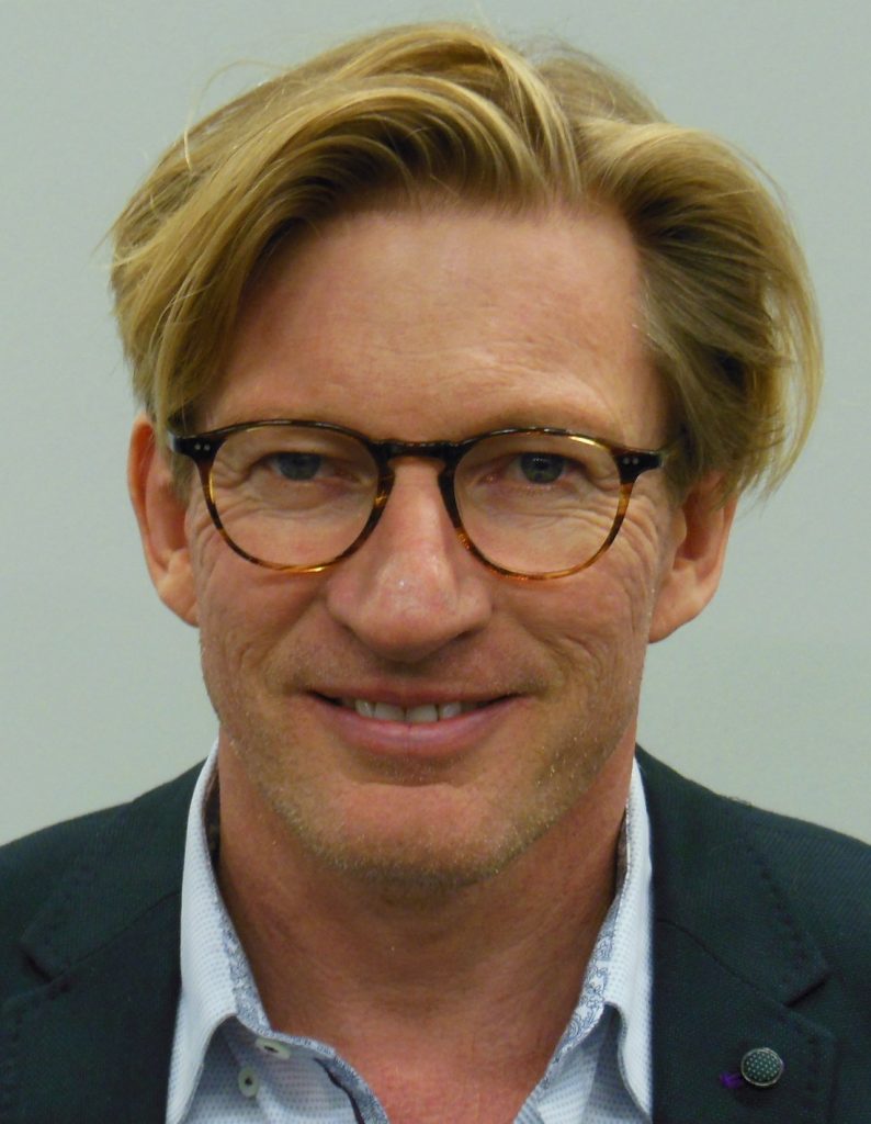 David Wenham, cheveux blonds coiffés en arrière et lunettes à monture écaille, sourit face à l'objectif, en veste sombre sur une chemise claire.