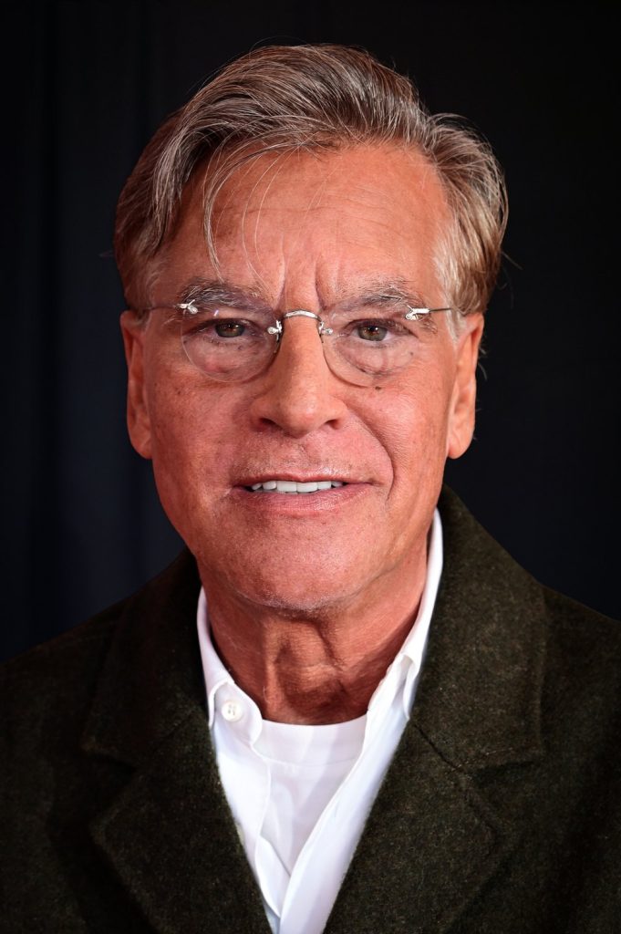 Aaron Sorkin, cheveux gris et lunettes, en veste sombre et chemise blanche, sourit devant un fond noir.