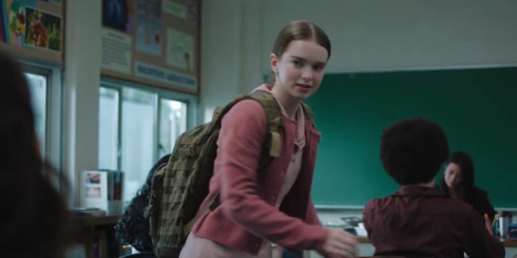 Une lycéenne en gilet rose, sac à dos sur l'épaule, entre dans une salle de classe où des élèves sont déjà assis.