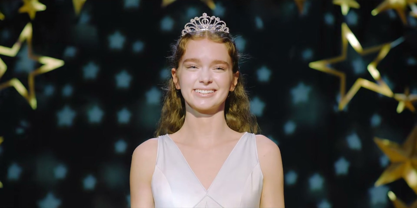 Une adolescente rousse en robe blanche, une tiare sur la tête, sourit devant un décor d'étoiles dorées.