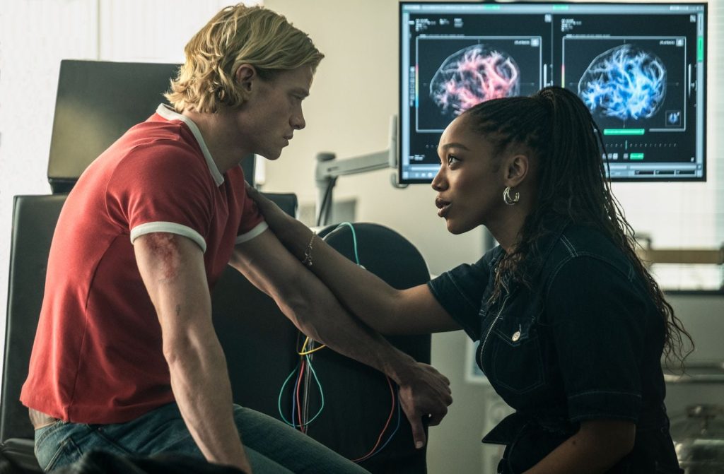 Naomi Ackie pose des électrodes sur le bras de Tom Rhys Harries, assis dans un laboratoire devant des écrans d’imagerie cérébrale.
