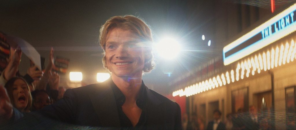 Tom Rhys Harries sourit sur un tapis rouge, devant une foule de photographes et l’enseigne d’un cinéma.