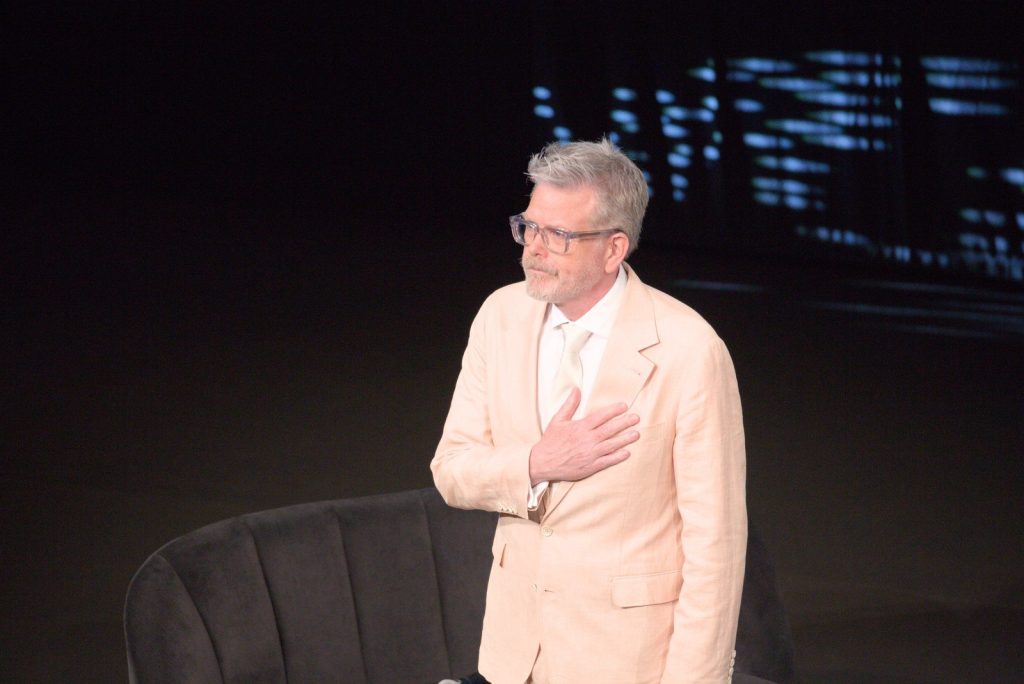 Christopher McQuarrie, en costume clair et lunettes, la main sur le cœur, debout devant un fauteuil sur une scène sombre.