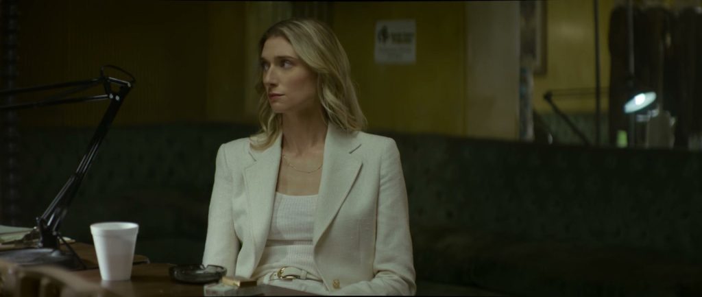 Elizabeth Debicki, en tailleur blanc, assise près d'un gobelet en polystyrène dans un bureau aux murs vert sombre.