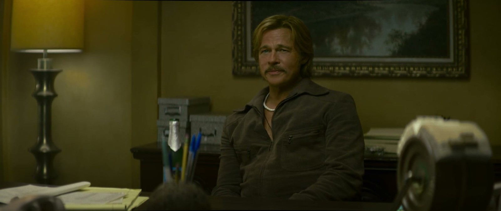 Brad Pitt, moustache et veste marron, assis dans un bureau éclairé par une lampe, devant un tableau encadré.