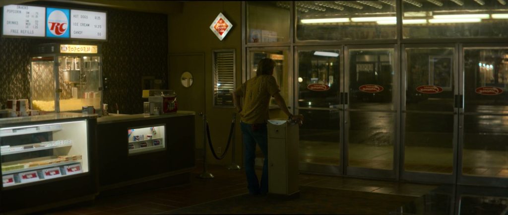 Un homme de dos devant les portes vitrées du hall d'un vieux cinéma, à côté du comptoir de confiserie et d'une enseigne RC Cola.