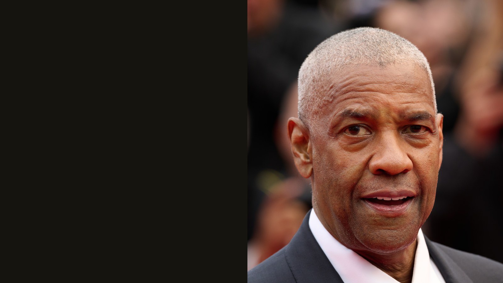 Denzel Washington, cheveux gris très courts, en costume sombre et chemise blanche, tourne la tête vers l'objectif, sur un tapis rouge.