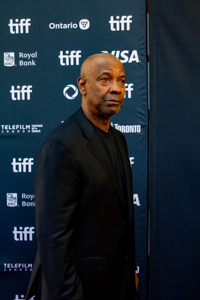 Denzel Washington, en costume noir, de profil, passe devant un mur de logos du festival de Toronto.