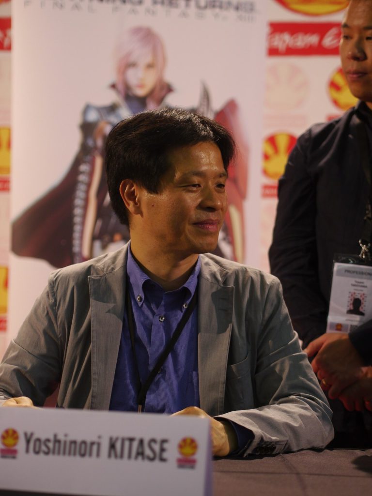 Yoshinori Kitase sourit, assis à une table de dédicace derrière un chevalet à son nom, devant une affiche de Lightning Returns: Final Fantasy XIII.