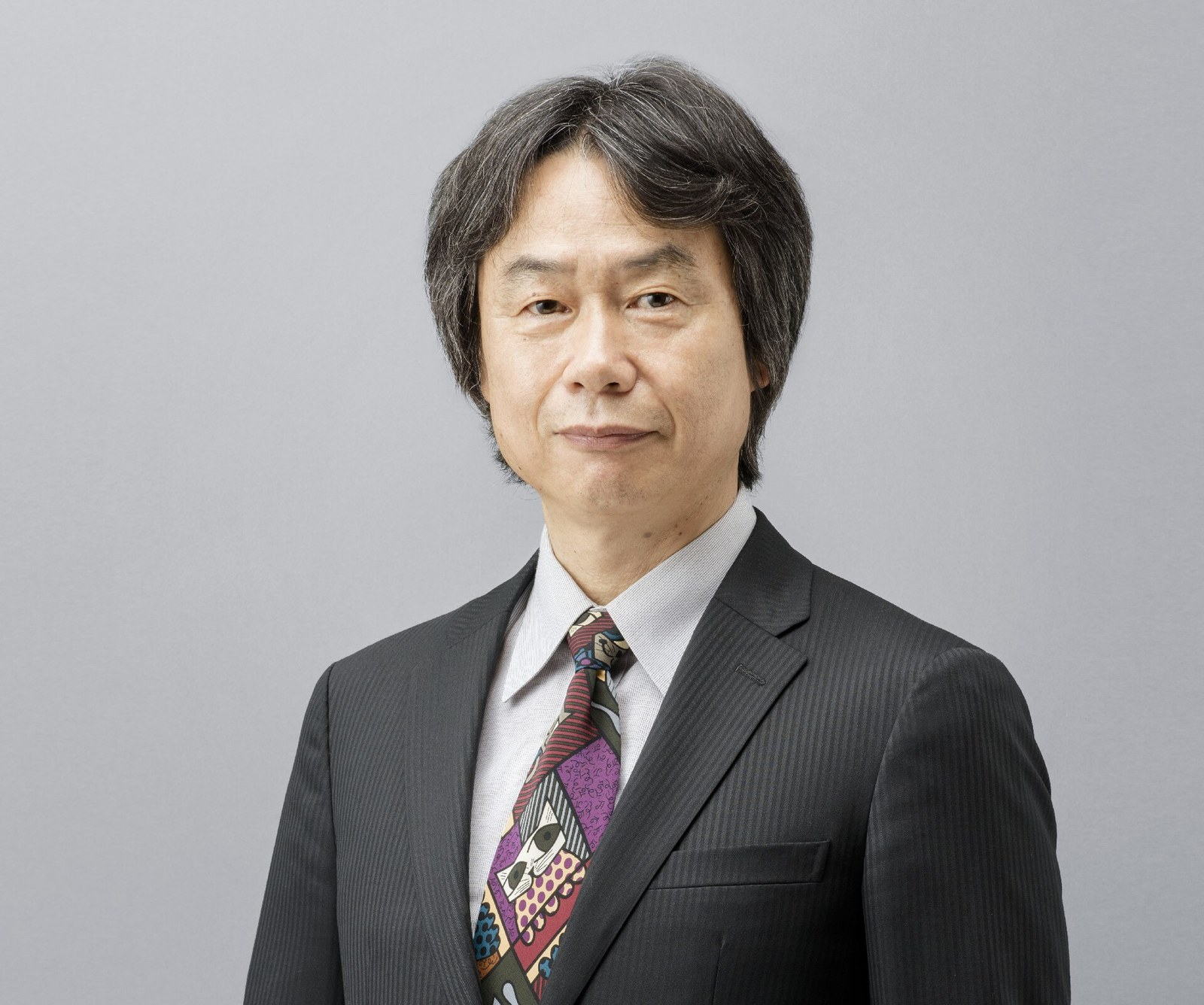 Shigeru Miyamoto en costume sombre et cravate colorée, devant un fond gris clair.