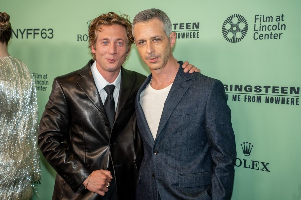 Deux hommes posent côte à côte devant un panneau vert de festival, l'un en veste de cuir noir, l'autre en costume bleu et tee-shirt blanc.
