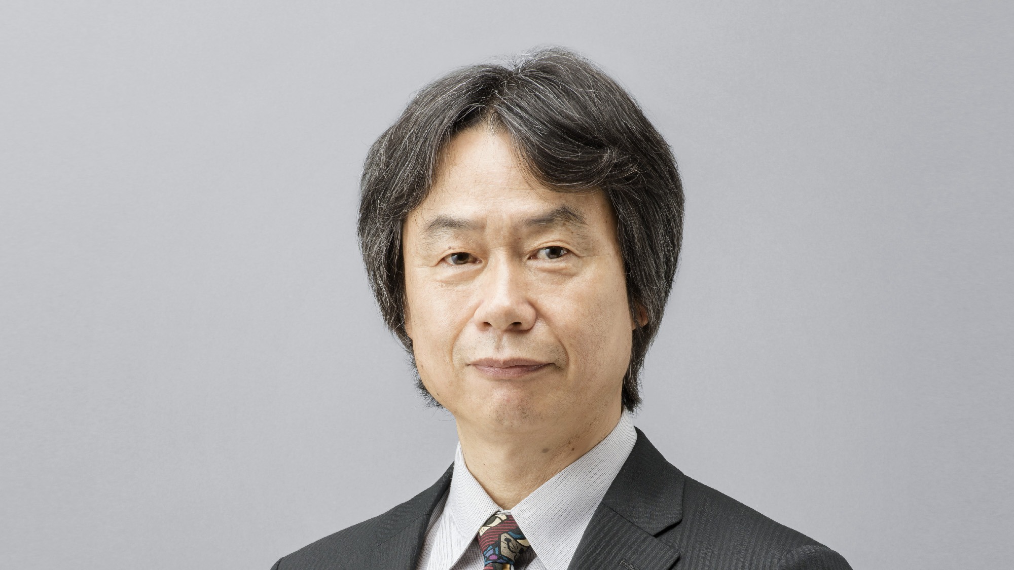 Shigeru Miyamoto en costume sombre et cravate à motifs, sur un fond gris clair.