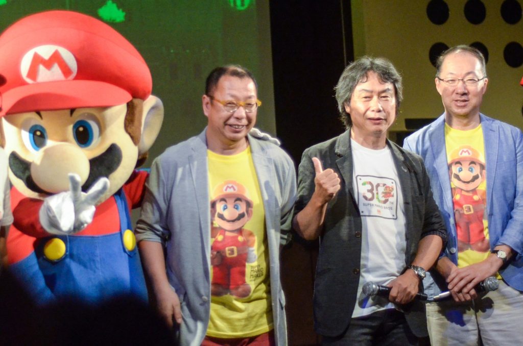 Trois hommes souriants sur une scène, à côté d'une mascotte de Mario. Celui du milieu, en tee-shirt blanc, lève le pouce.