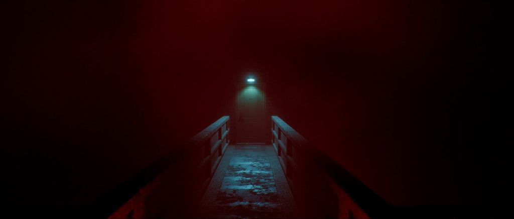 Un couloir étroit baigné de lumière rouge mène à une porte éclairée par une petite lampe.