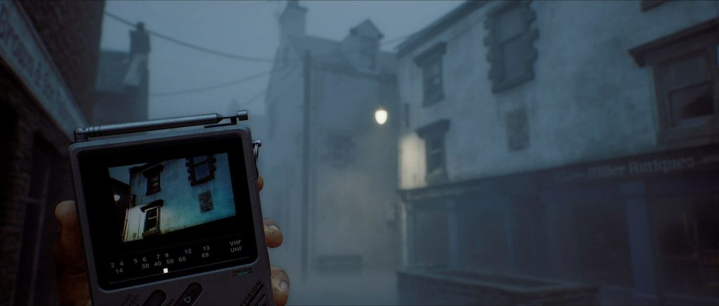 Une main tient une petite télévision portable dont l'écran montre une façade, dans une rue brumeuse bordée de maisons de pierre.