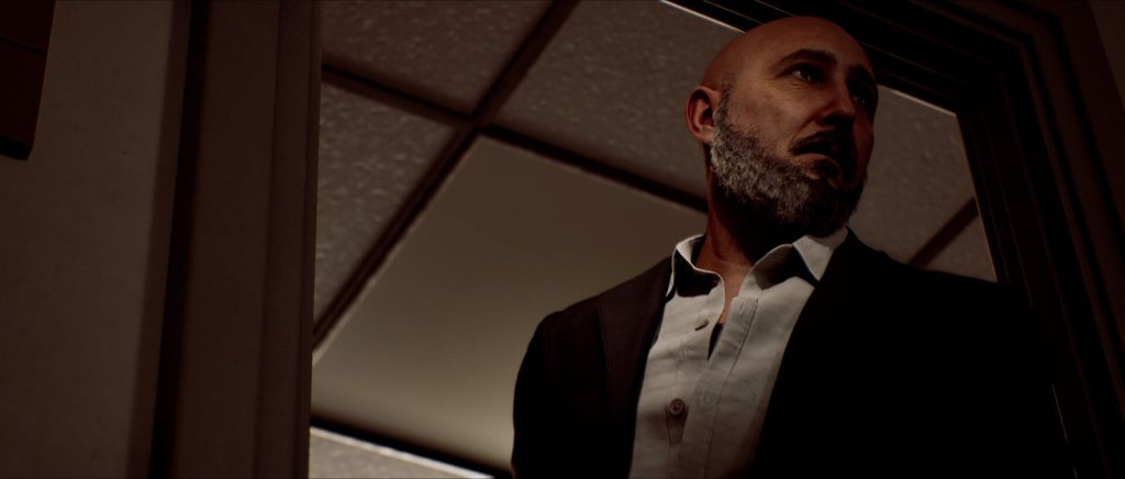 Un homme chauve à la barbe grise, en veste sombre et chemise blanche, vu en contre-plongée près d'une fenêtre.