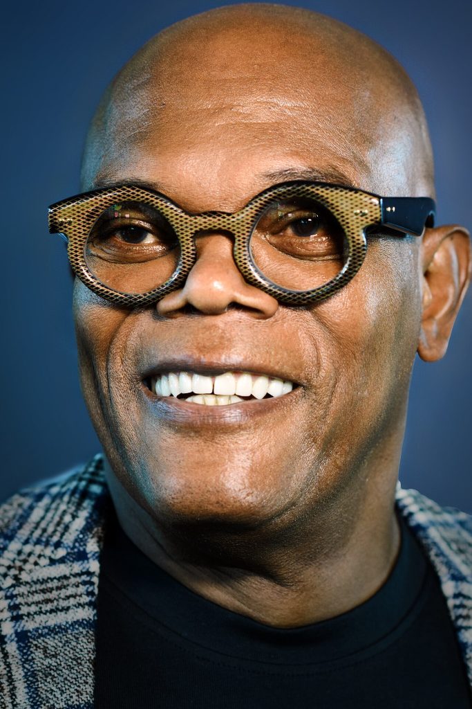 Portrait souriant d’un homme au crâne rasé, lunettes rondes à monture travaillée, veste à carreaux sur un col noir, devant un fond bleu.