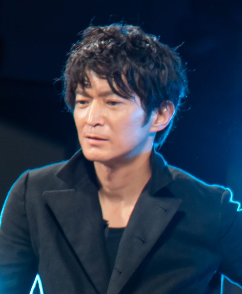 Portrait d’un homme aux cheveux noirs en bataille, veste sombre sur un col roulé, de trois quarts, devant un fond bleu.