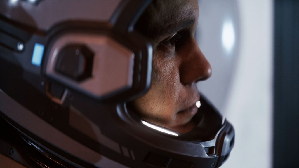 Gros plan sur le visage d'un homme derrière la visière de son casque spatial, éclairé de blanc.