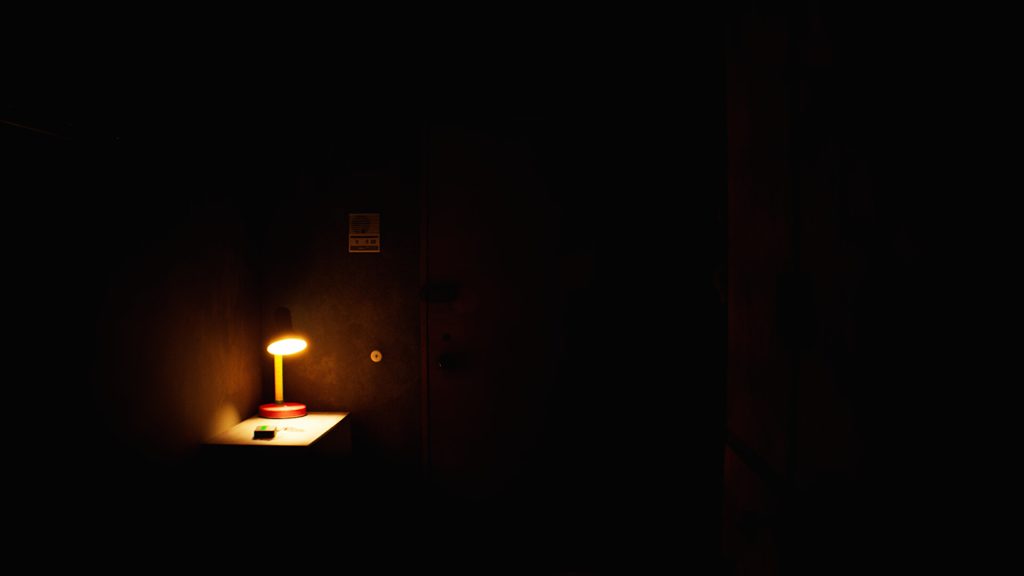 Une lampe de bureau allumée éclaire une feuille posée sur une tablette, seul point de lumière dans une pièce noire.